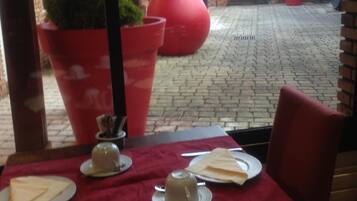 Un área de comedor al aire libre con mesas preparadas para dos, plantas grandes en macetas y un camino de ladrillo que conduce a un edificio.