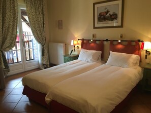 A double bed with white linens in a room with a radiator, a framed picture on the wall, and a window with curtains.