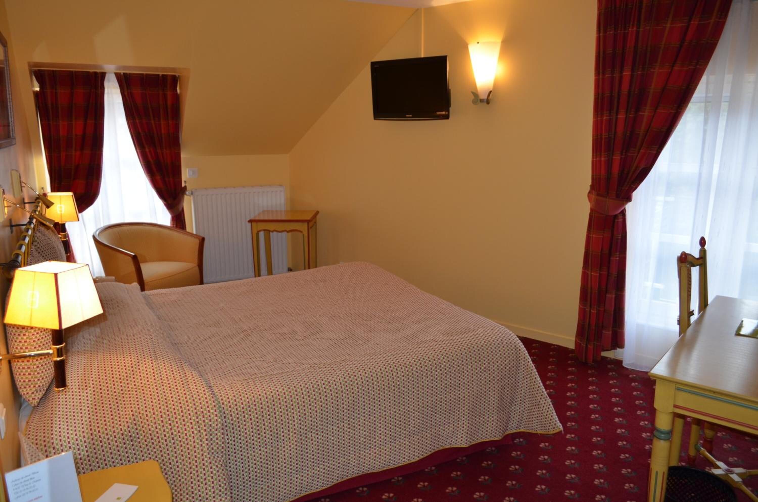 A hotel room with a bed, a chair, a small table, a lamp, a TV, and a window with curtains.