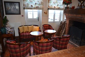 A cozy living room with a fireplace, plaid chairs, a sofa with cushions, and a window with floral curtains.