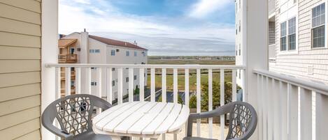 A balcony with a white table and two chairs, overlooking a residential area with houses and a clear sky.