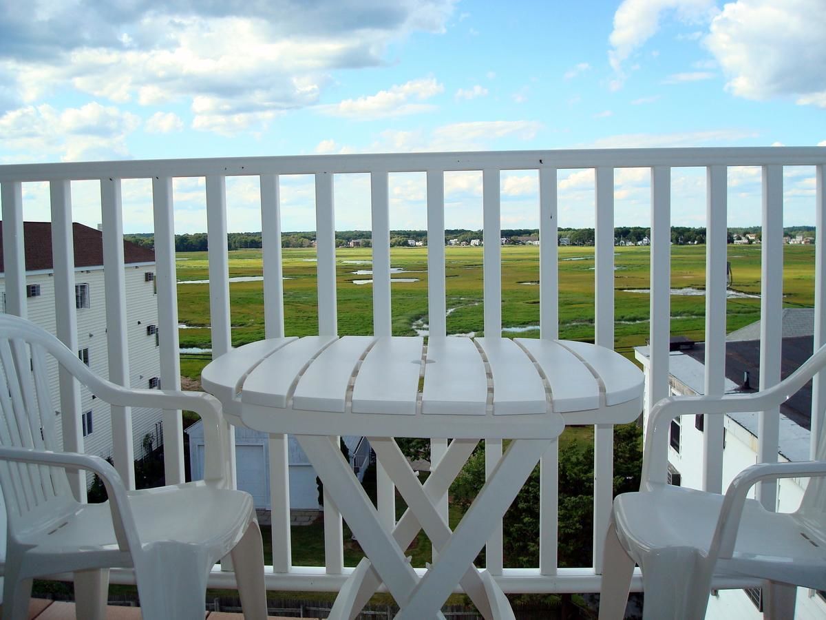 A balcony with a white table and chairs, overlooking a green landscape with buildings in the distance.