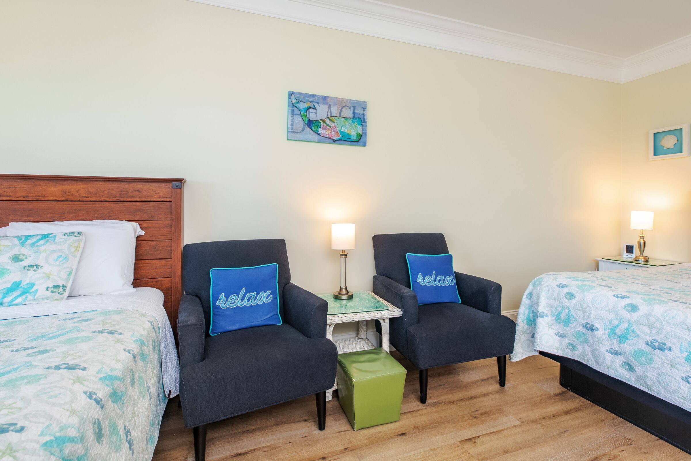 A hotel room with two beds, a wooden headboard, two armchairs with 'relax' pillows, a small table with a lamp, and a wall art piece.
