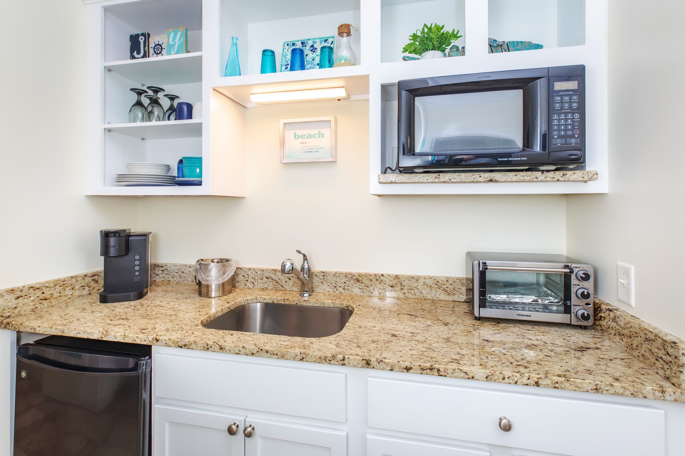 A kitchen with a granite countertop, a stainless steel sink, a black microwave, a toaster oven, and a coffee maker.