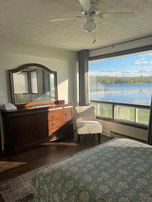 A bedroom with a large bed, a dresser, a chair, a ceiling fan, and a window with a lake view. - Welcome Inn (Papineau-Cameron)