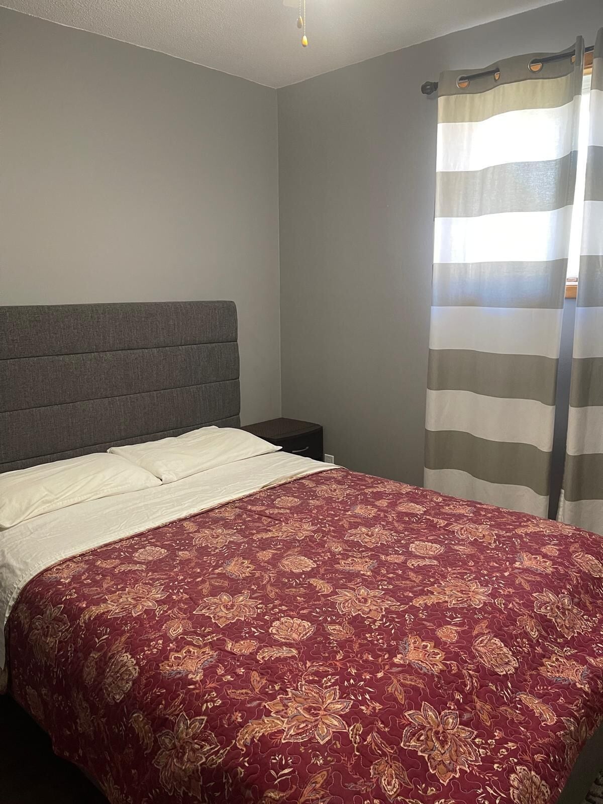 Un dormitorio con cama, cabecera gris y cortina a rayas.