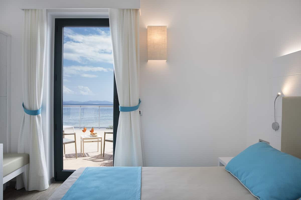 A hotel room with a bed, a view of the sea, and a balcony with chairs.