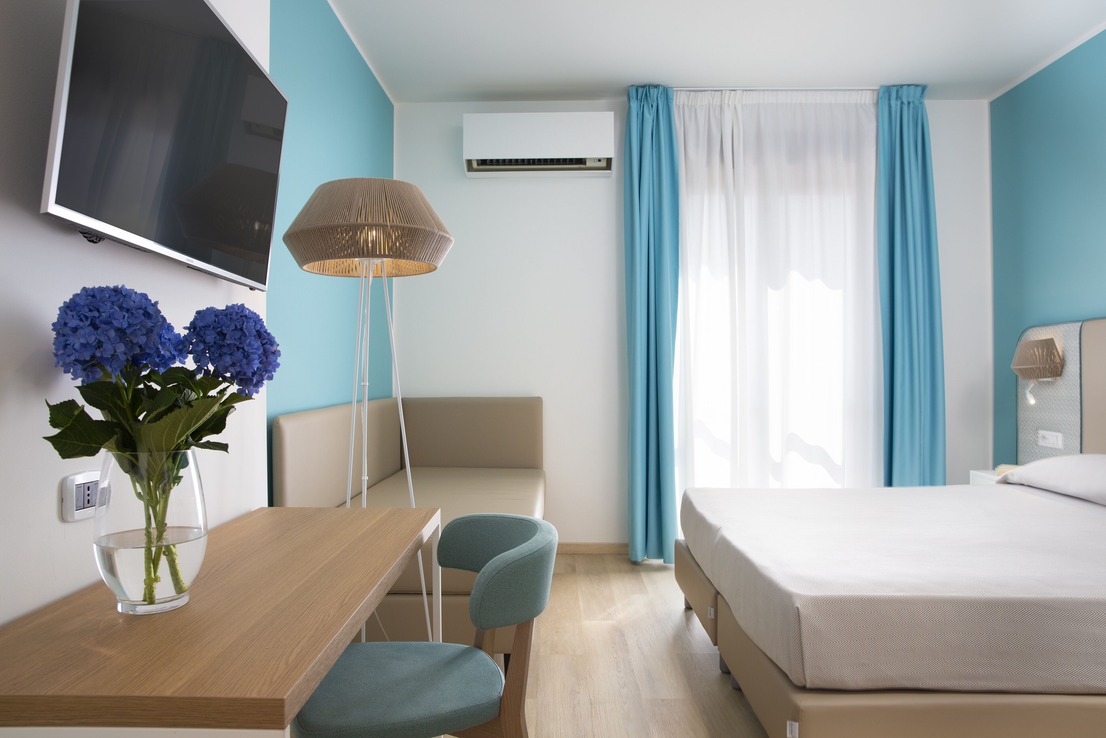 A hotel room with a bed, a desk with a chair, a TV mounted on the wall, an air conditioning unit, and a window with curtains.