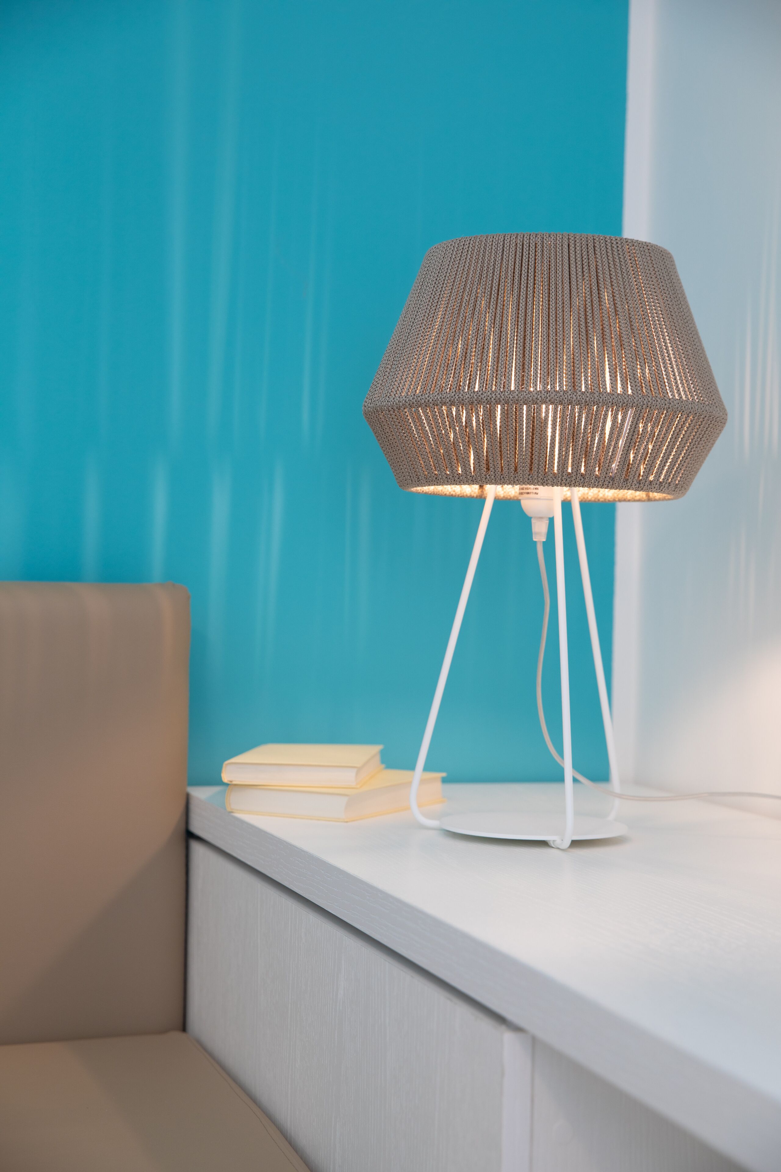 A modern lamp with a woven shade on a white table against a teal wall.