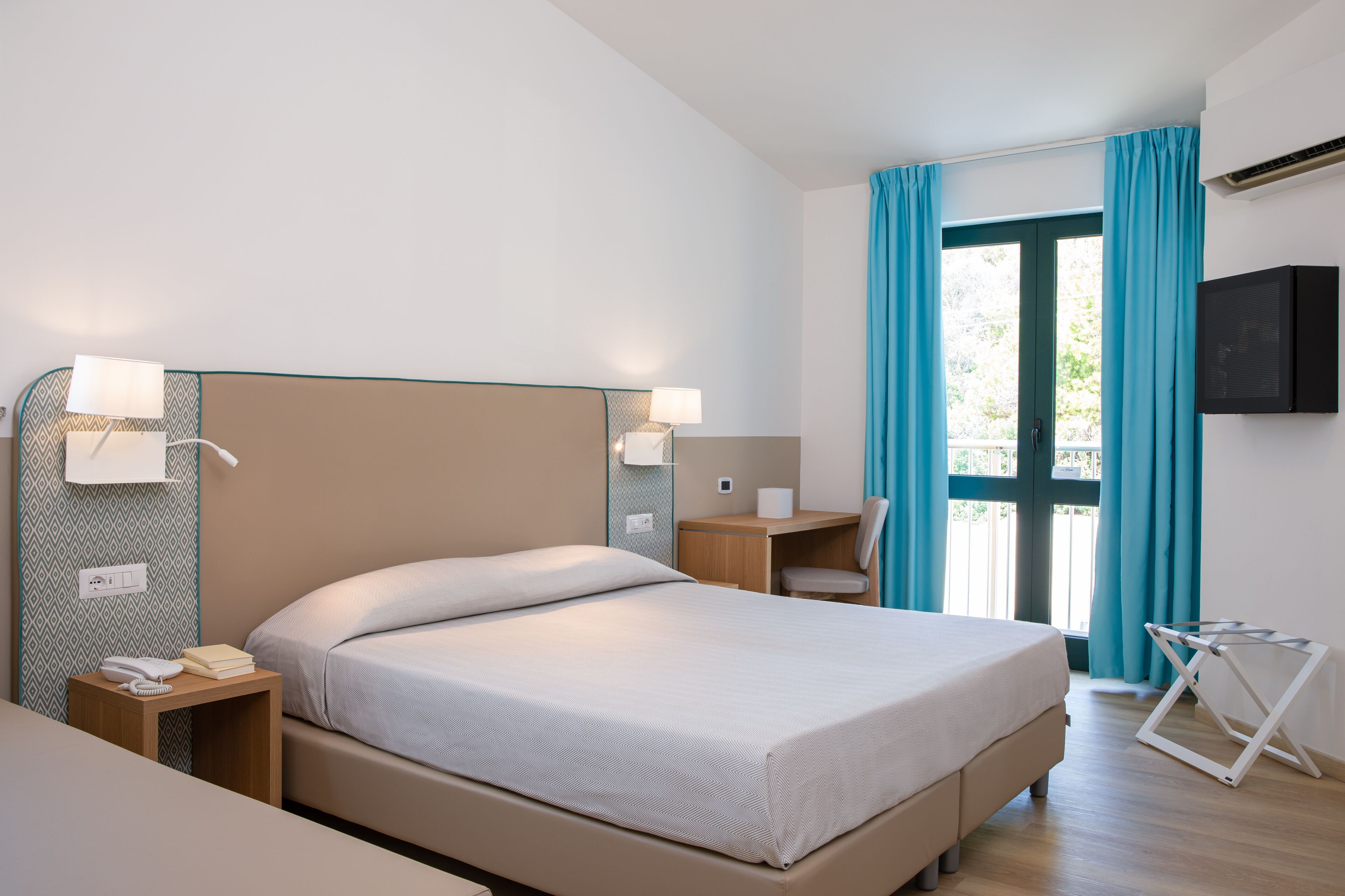 Une chambre d’hôtel comprenant un lit, des tables de chevet, un bureau, une chaise, une télévision et une fenêtre avec des rideaux.