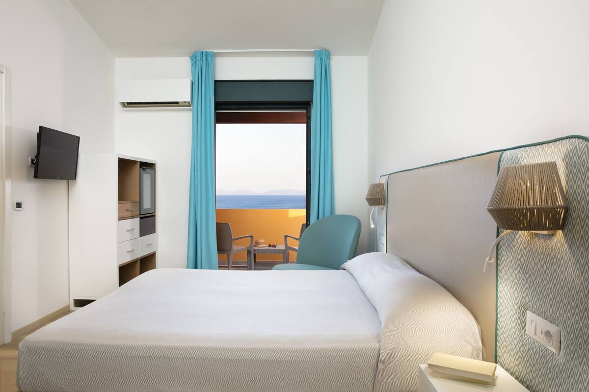 A hotel room with a large bed, a TV mounted on the wall, a desk with a chair, and a view of the outdoors through a window with blue curtains.