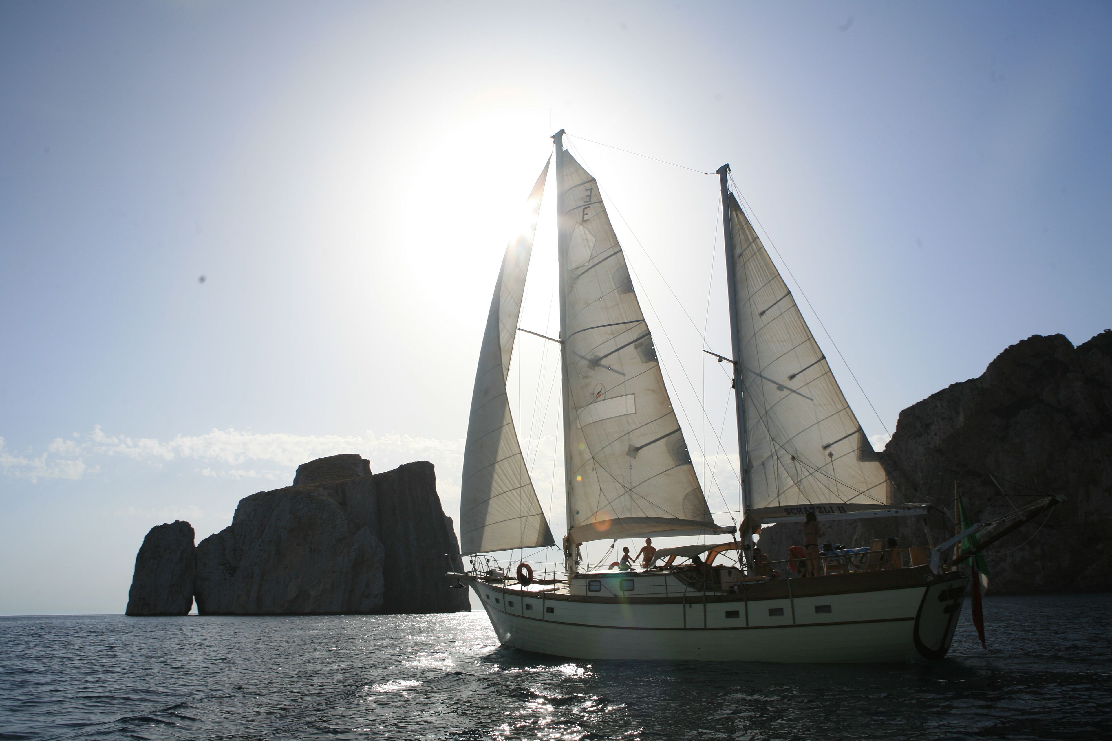 Ein Segelboot mit mehreren Segeln, vor felsigen Klippen vor Anker.