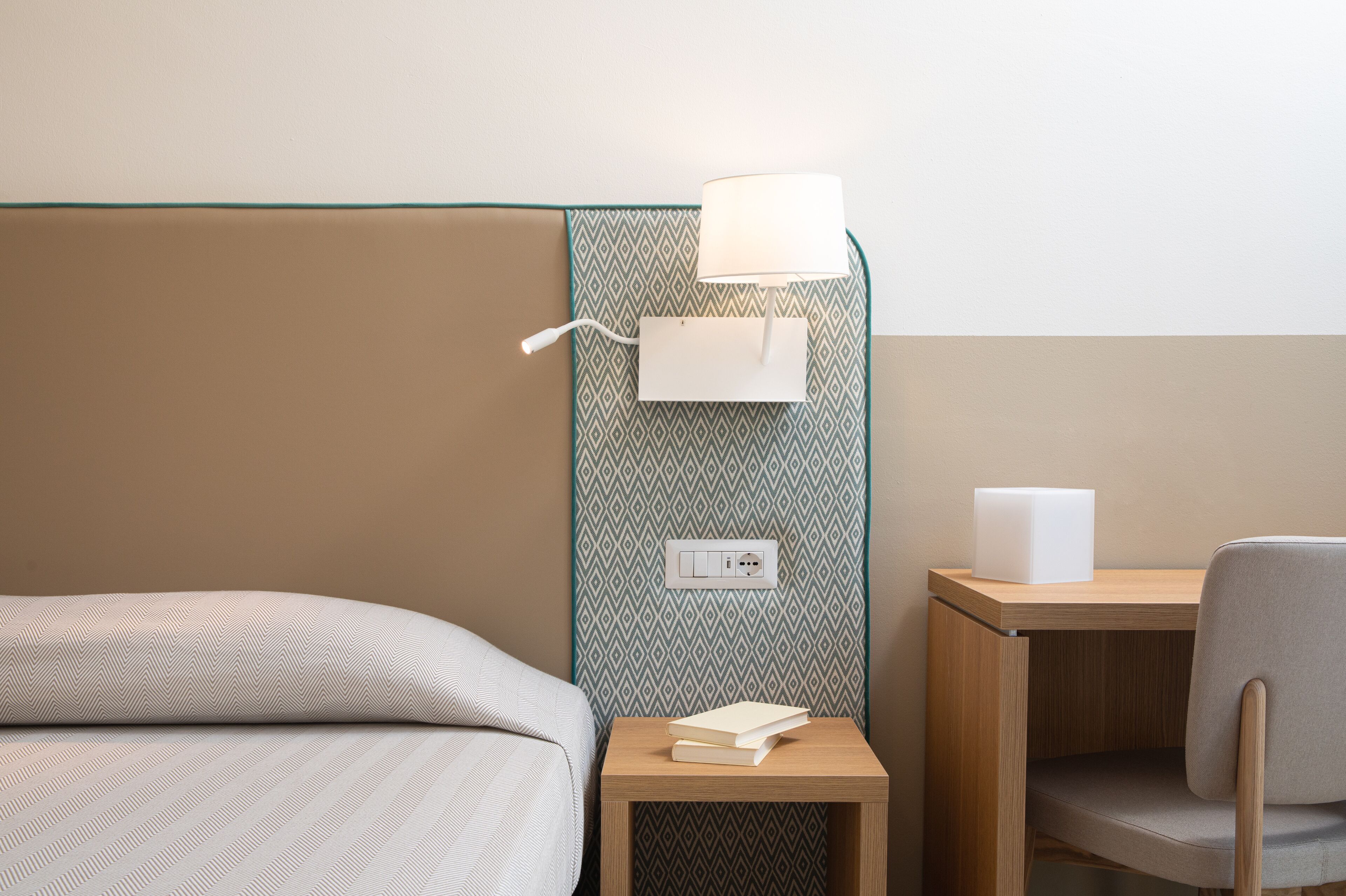 A hotel room with a bed, a bedside table with a lamp, a small desk with a chair, and a patterned wall.