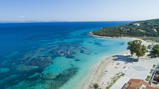 An aerial view of a coastal area with clear blue waters, a sandy beach, and buildings on a hillside.