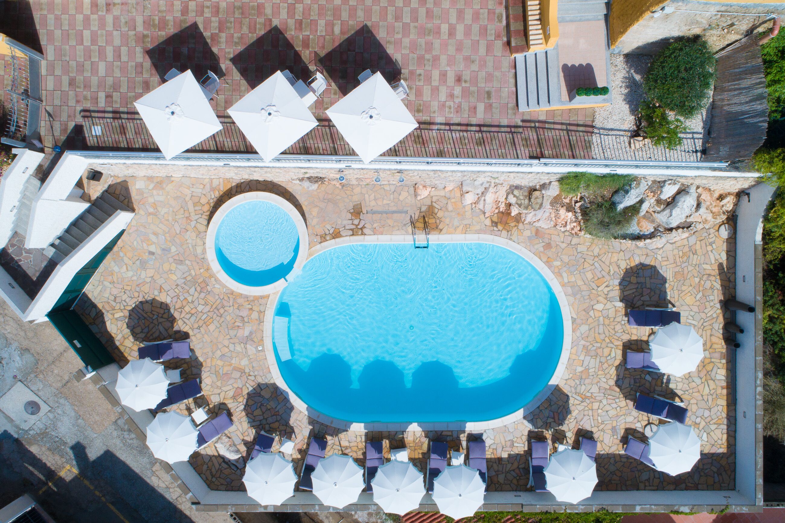 An aerial view of a swimming pool area with multiple lounge chairs and umbrellas, surrounded by a stone-tiled deck.