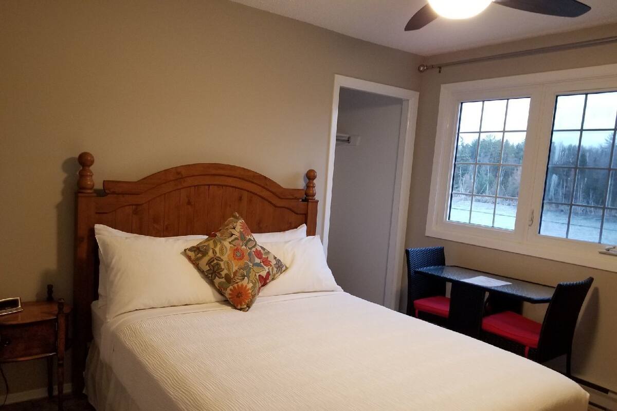 Een slaapkamer met een houten hoofdbord, een bed met witte lakens, een bloemenkussen, een nachtkastje, een bureau met stoelen en een raam met uitzicht op bomen.