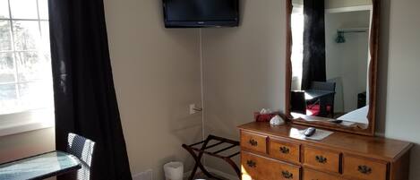 Una habitación de hotel con cama, cómoda, televisor montado en la pared, silla y ventana con cortinas.
