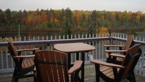 A wooden deck with a table and four Adirondack chairs, overlooking a lake with autumn-colored trees. - Bee 'n Bee Bed n' Breakfast (Lambton Shores)