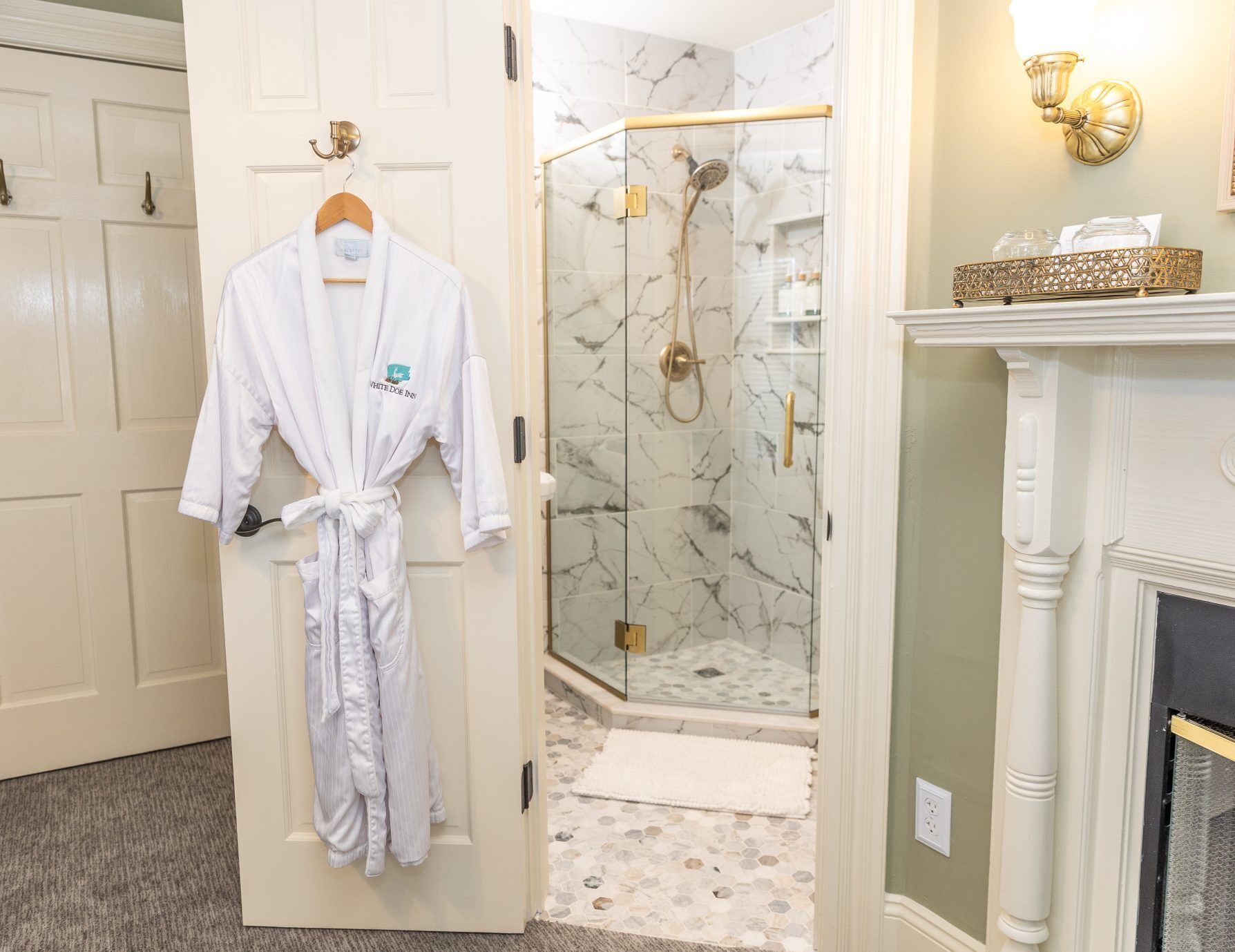 A hotel bathroom with a glass-enclosed shower, marble walls, a white robe hanging on a door, and a fireplace.