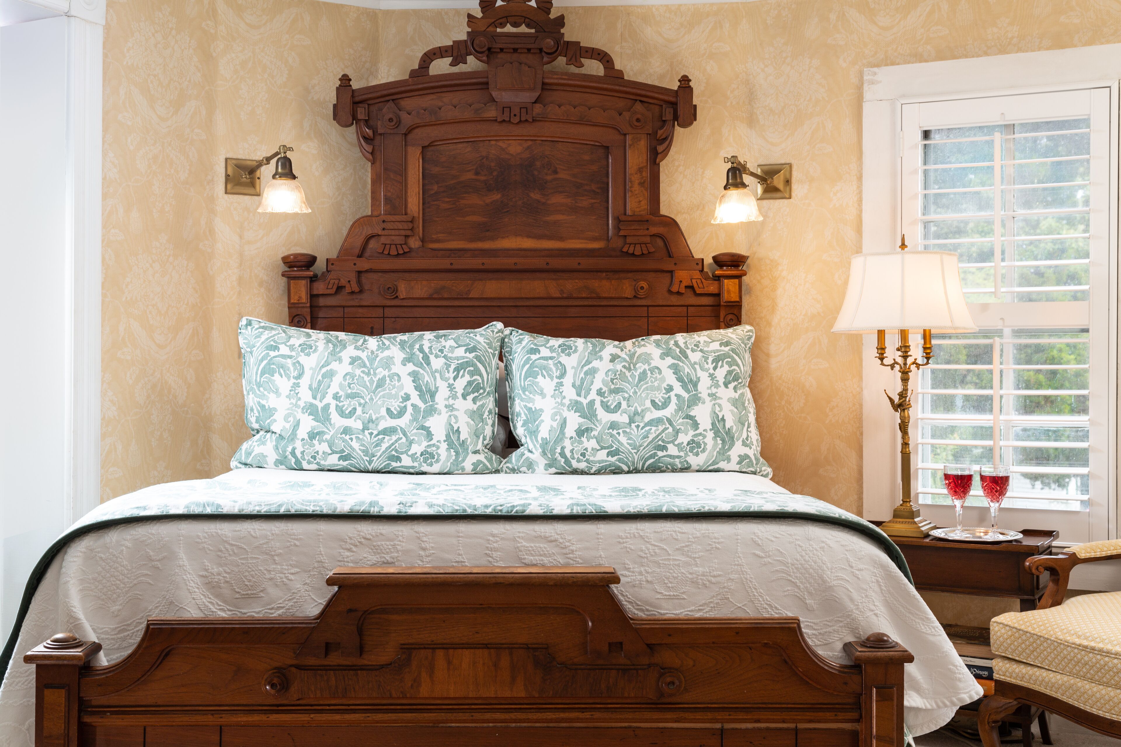 A wooden bed with a headboard, two wall-mounted lamps, and a bedside table with a lamp and two glasses.