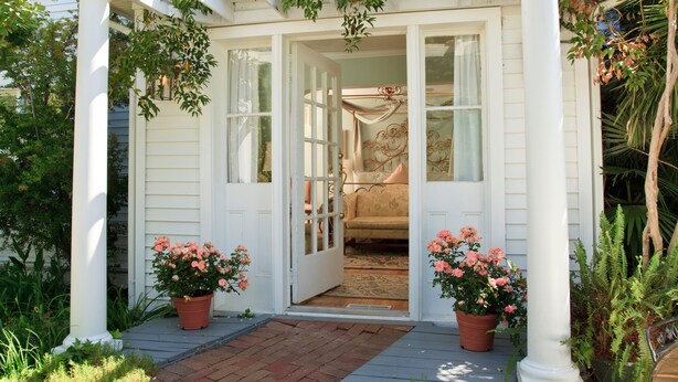 Een wit terras met een bankje, plantenpotten en uitzicht op een kamer met een bed en gordijnen.