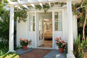 A white porch with a bench, potted plants, and a view into a room with a bed and curtains.