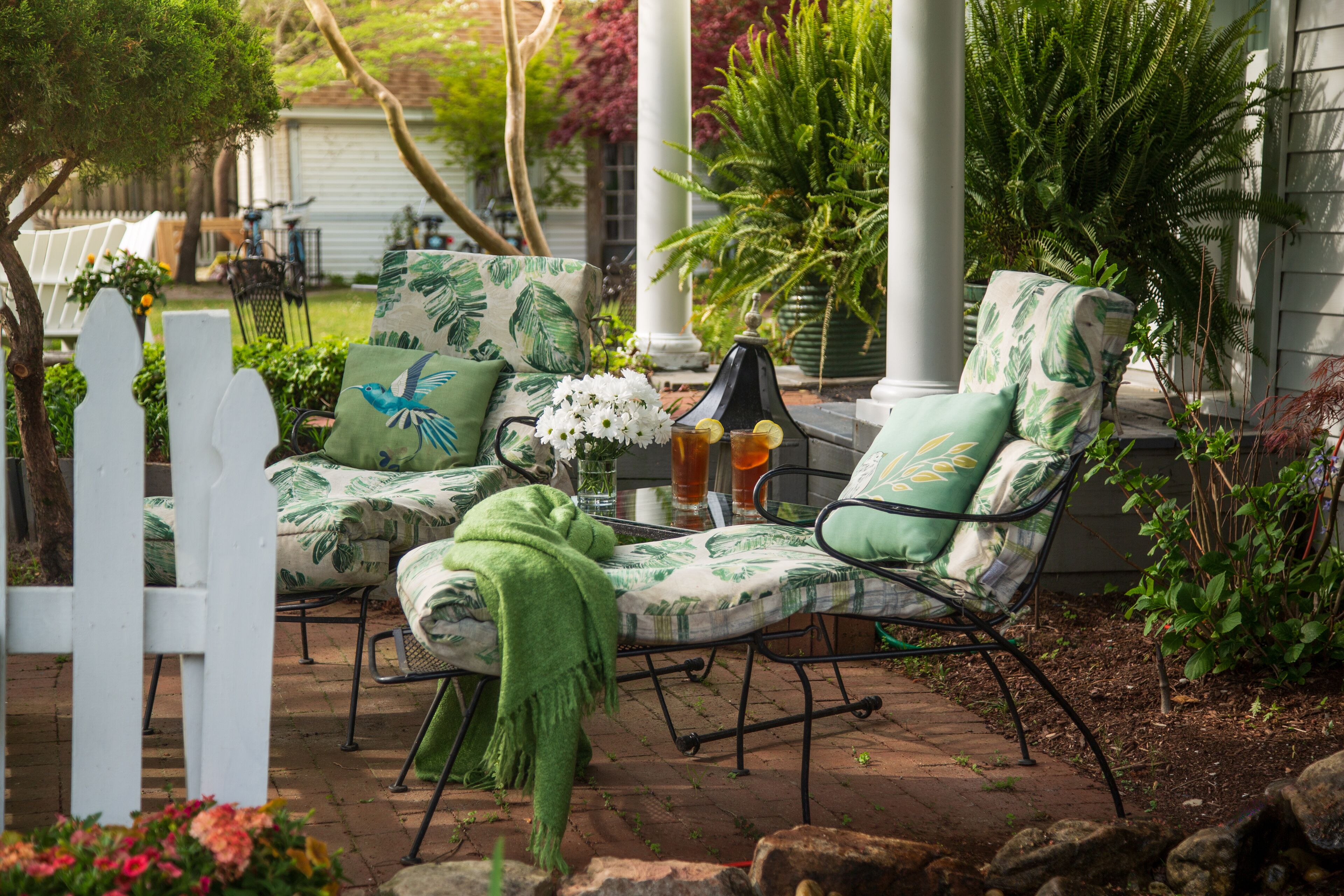 Un patio con dos sillones estampados en verde, una pequeña mesa con bebidas y una valla de piquetes blanca.