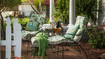 Un patio con dos sillones estampados en verde, una pequeña mesa con bebidas y una valla de piquetes blanca.