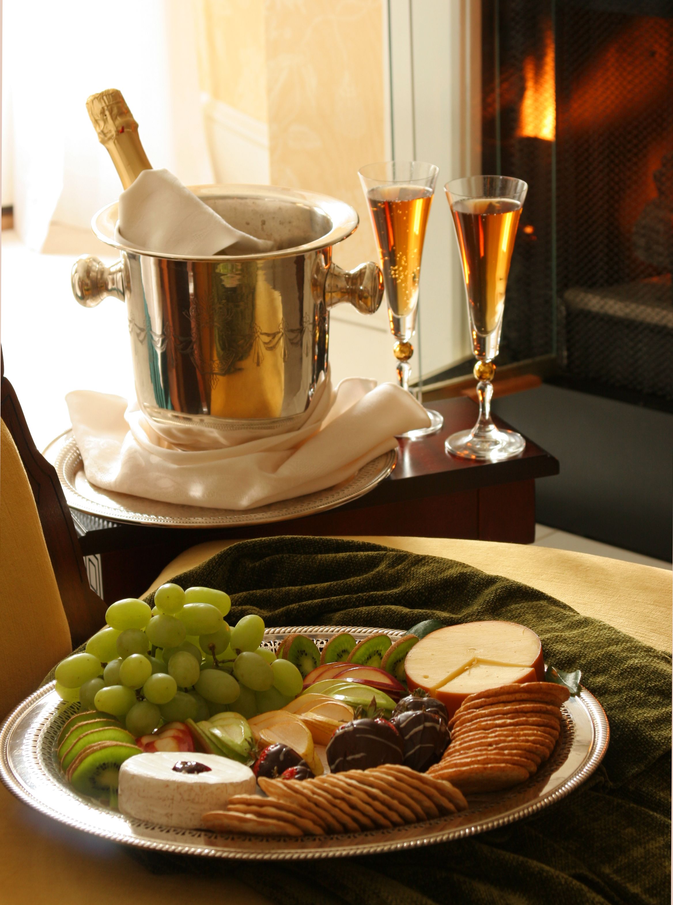 A room with a fireplace, a table with a bottle of champagne, two glasses of champagne, and a plate of cheese and fruits.