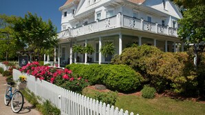 Een groot, wit koloniaal herenhuis met een veranda, omgeven door weelderige begroeiing en een witte picket fence.