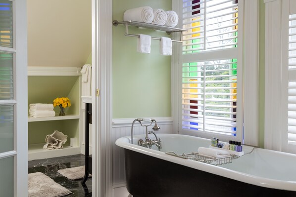 A bathroom with a freestanding bathtub, a towel rack with folded towels, a shelf with towels and a vase of flowers, and a window with blinds.
