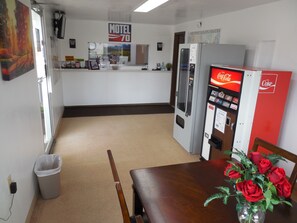 A motel lobby with a Coca-Cola vending machine, a refrigerator, a trash can, and a table with a vase of roses. - Motel 70 (High Hill)