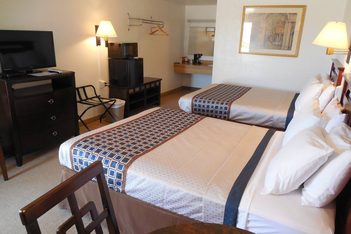 A hotel room with two beds, a desk with a TV, a chair, a small table, and a lamp.