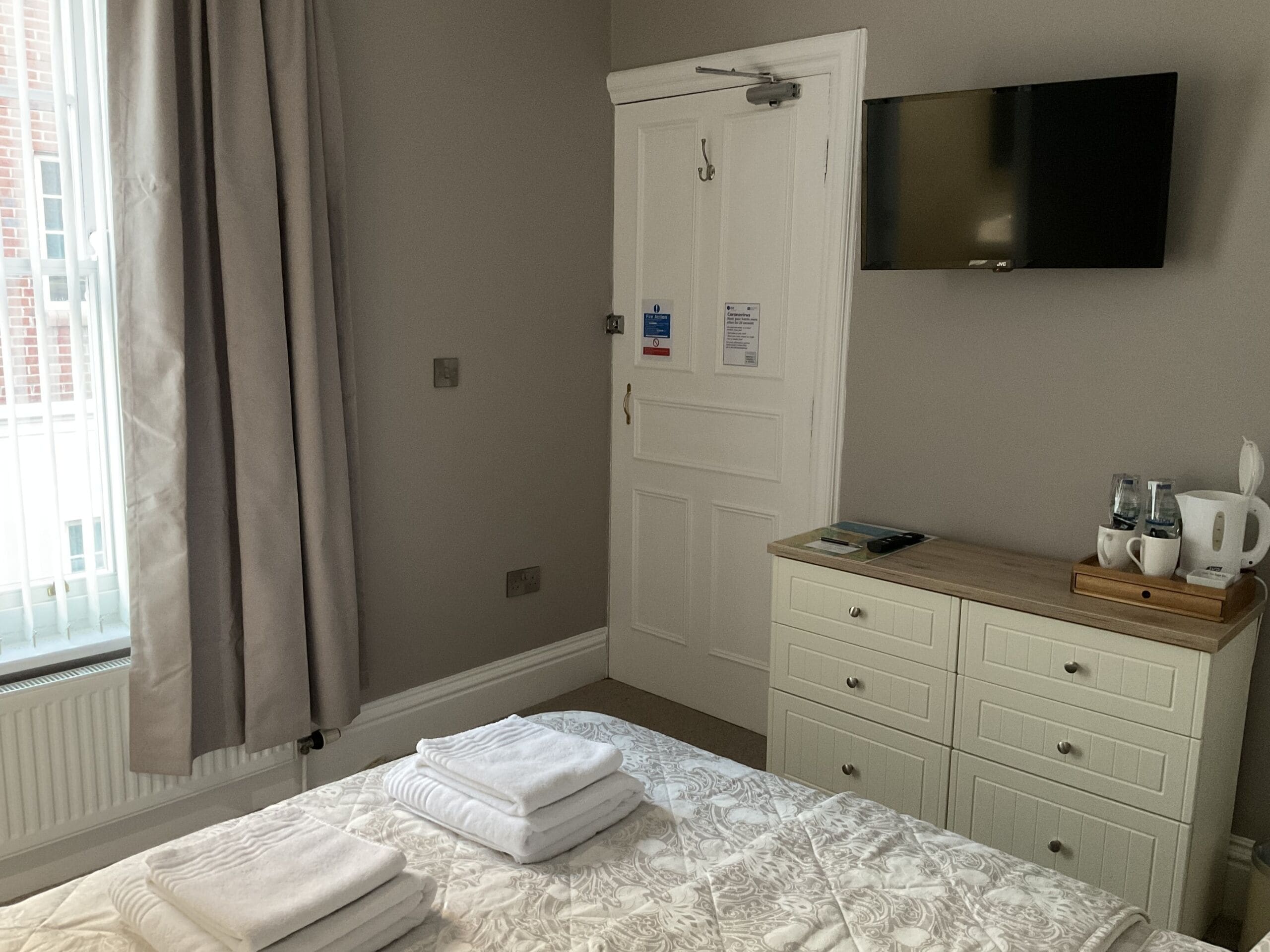 A hotel room with a bed, a TV mounted on the wall, a dresser with toiletries, and a window with curtains.