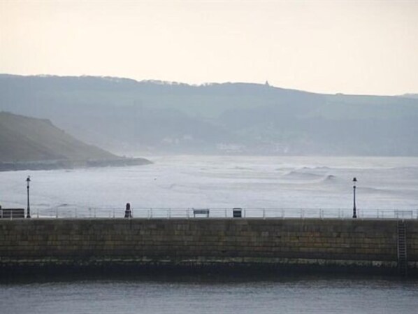 A coastal scene with a stone pier extending into the sea, a person standing on the pier, and a hill in the background. - Glenora Guest House (Whitby)