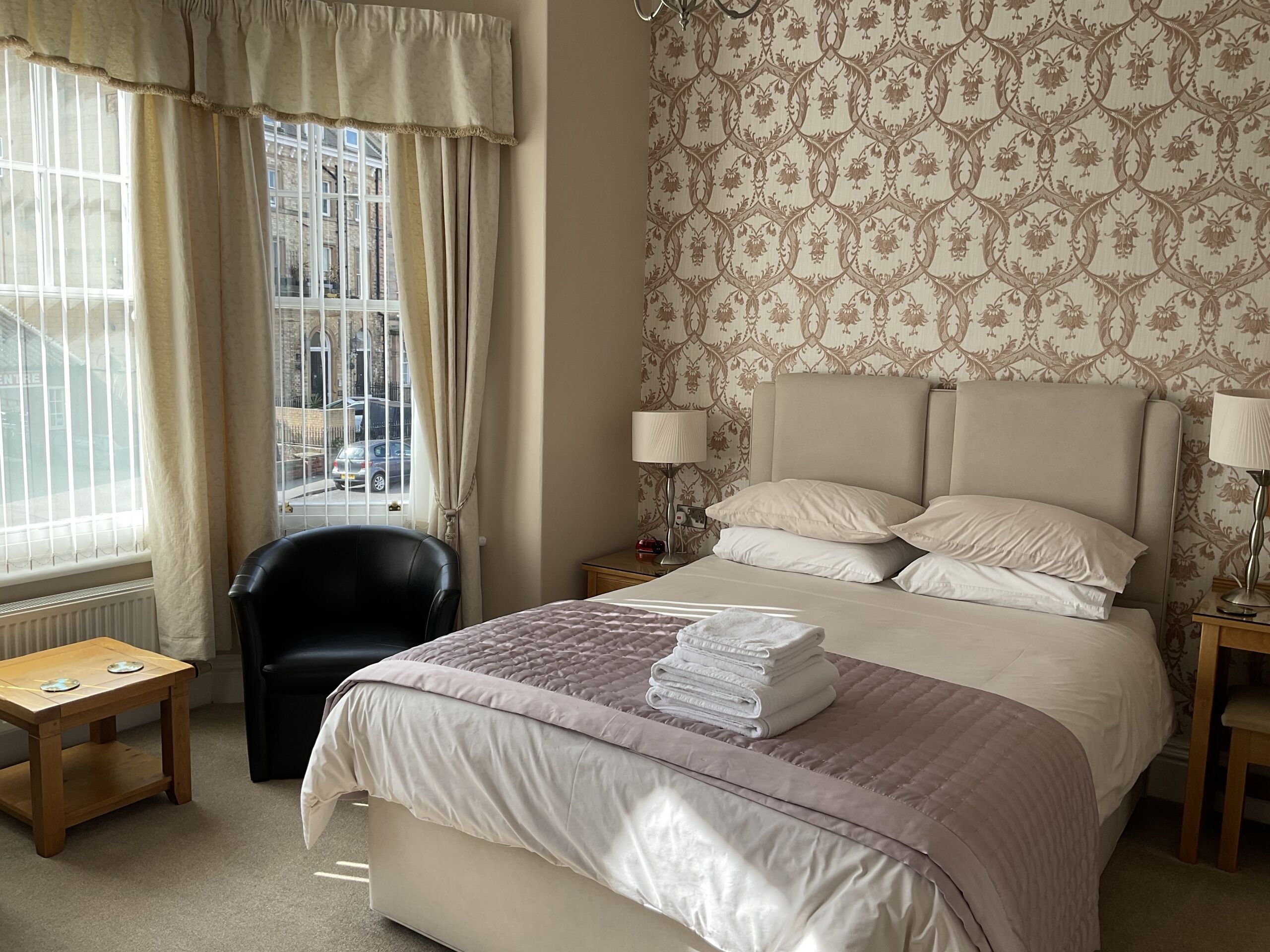 A hotel room with a large bed, a chair, a nightstand, a lamp, and a view of the street through the window.
