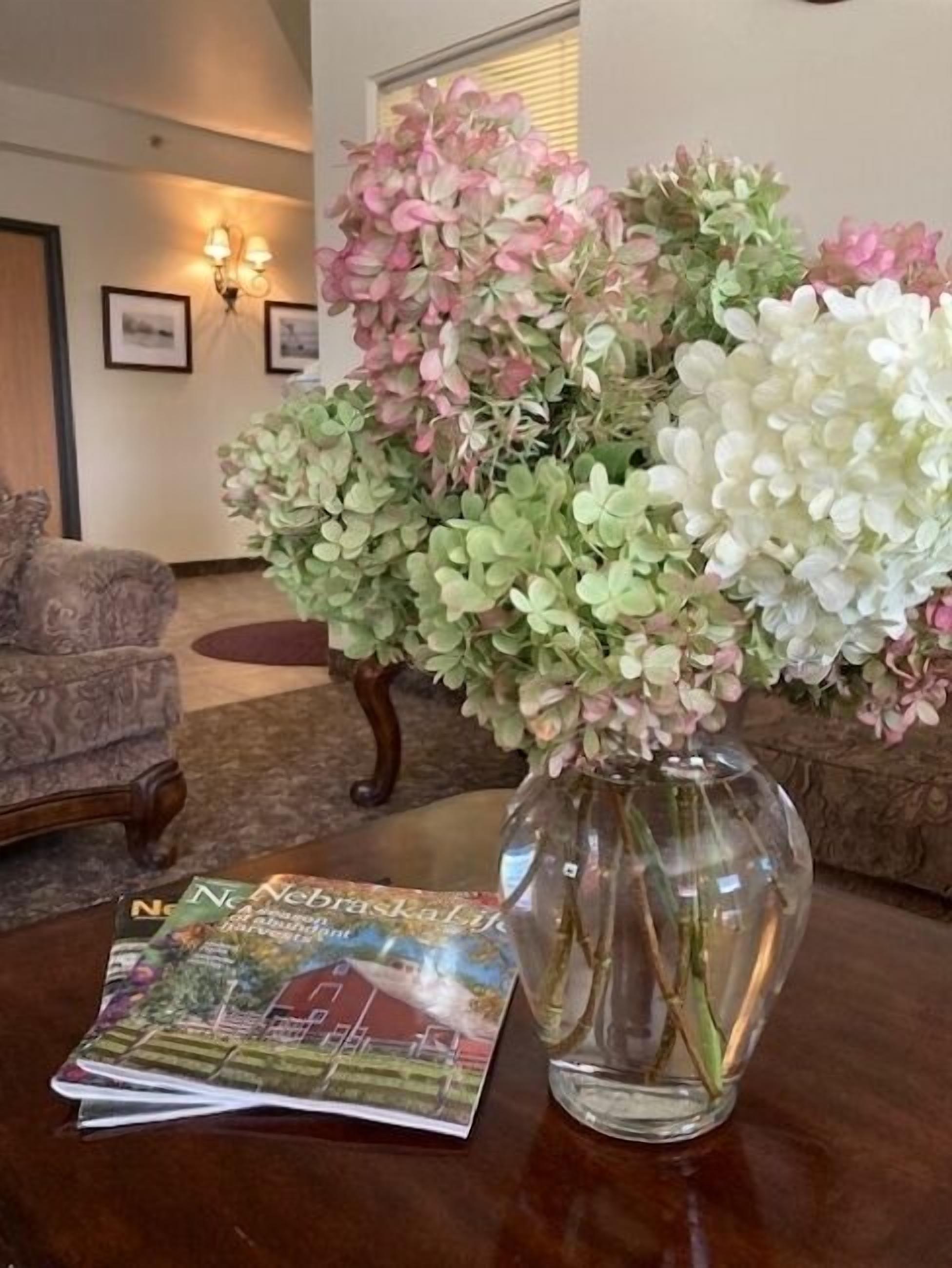 A clear glass vase with pink and white hydrangea flowers on a wooden table, next to a magazine.