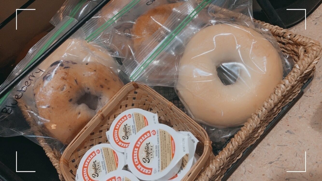 A basket containing various packaged food items, including a bagel, a blueberry bagel, and cheese spread.