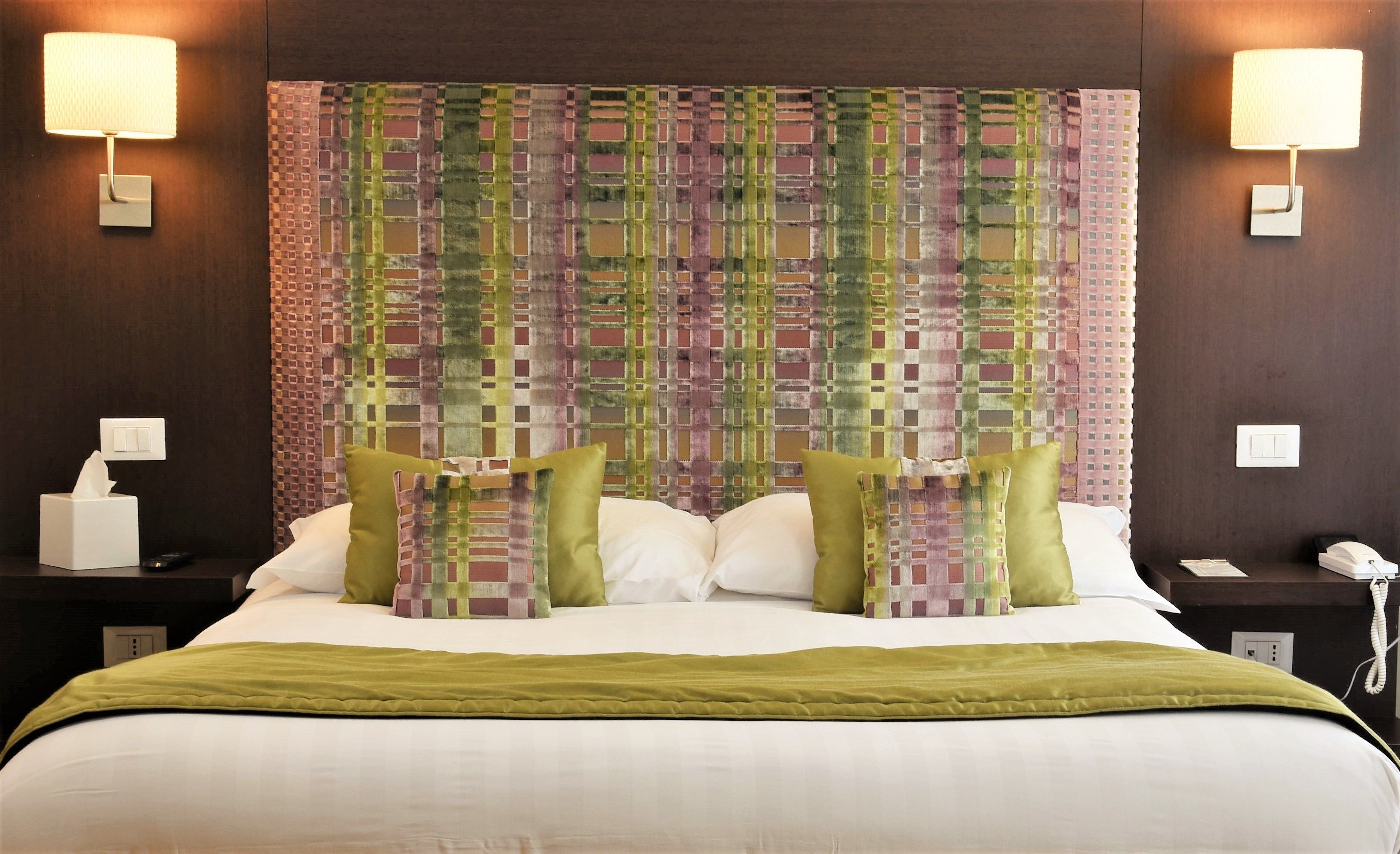 Ein Hotelzimmer mit einem großen Bett, zwei Nachttischlampen, einem gemusterten Kopfteil und einer grünen Tagesdecke.