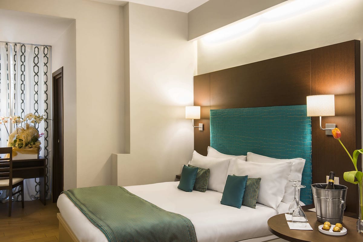 A hotel room with a bed, bedside table, lamp, and a vase with flowers.