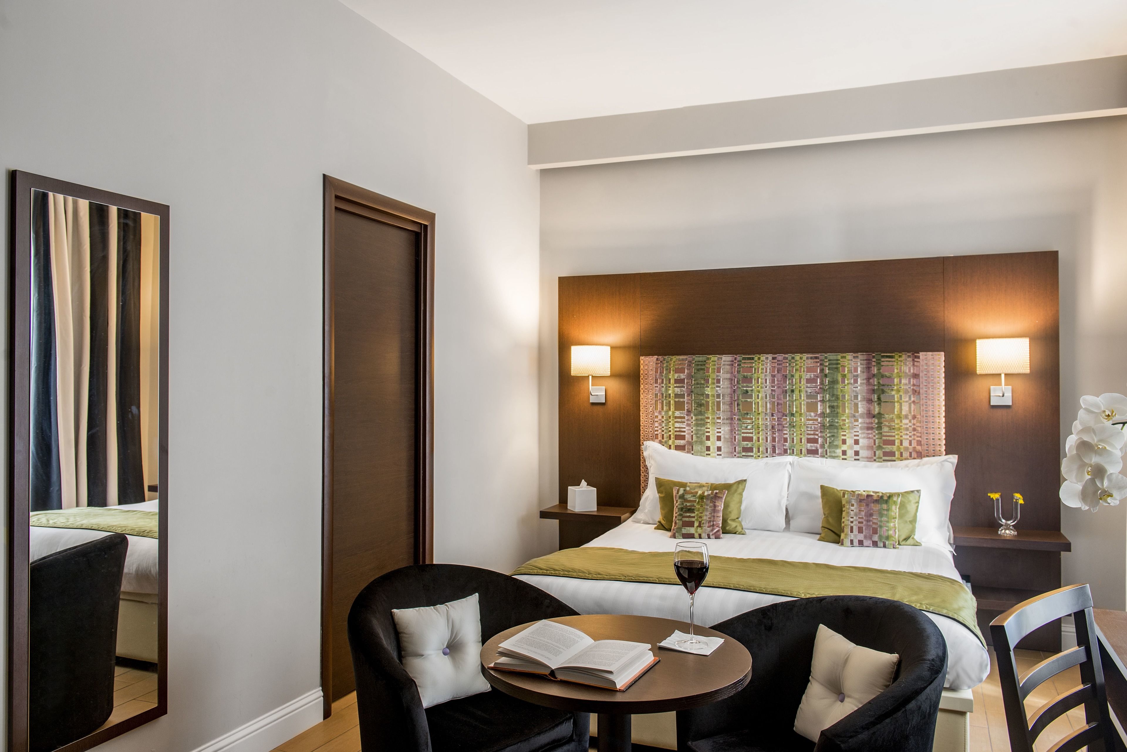 Una habitación de hotel con una cama grande, dos mesitas de noche, una mesa redonda con una silla, un espejo y una ventana.