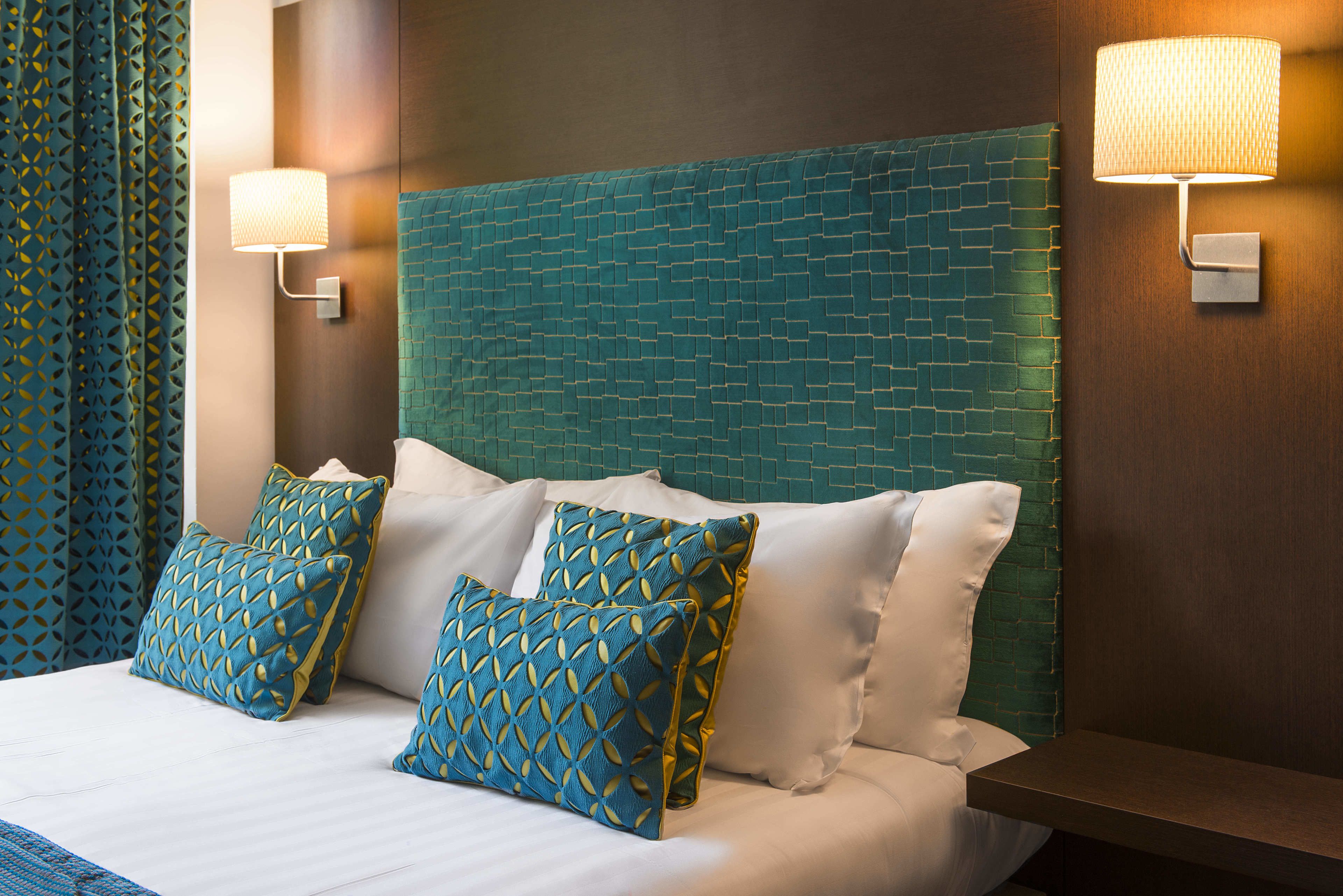 Habitación de hotel con cabecera de ladrillo, dos lámparas de pared y una cama con ropa blanca y almohadas decorativas.