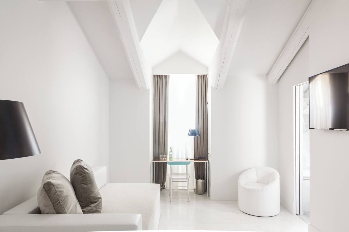 Habitació d'hotel moderna amb un sofà blanc, un escriptori petit amb una làmpada, un televisor fixat a la paret i una vista per una finestra amb cortines.