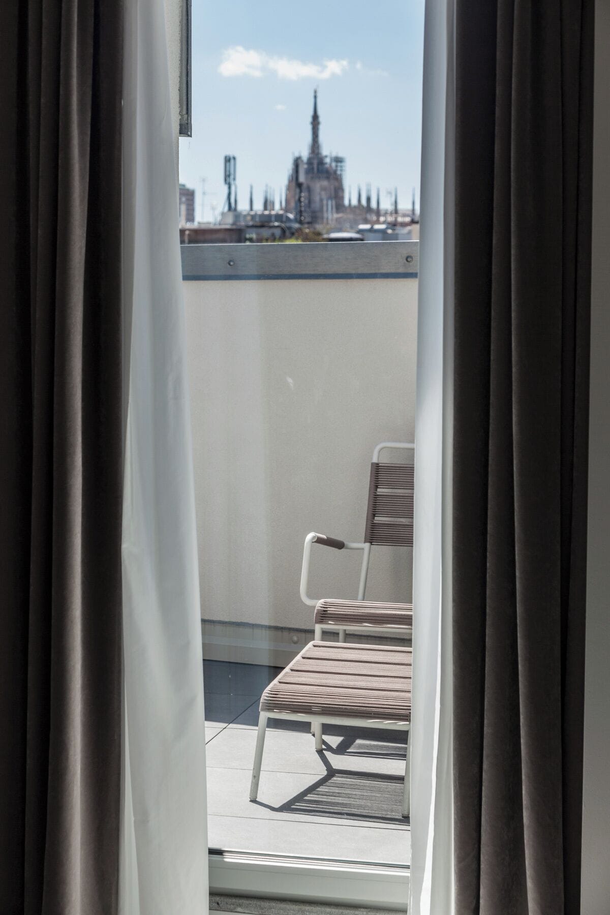 Ein Balkon mit einem Stuhl, Vorhängen und Blick auf eine Stadtlandschaft, darunter ein hohes Gebäude.