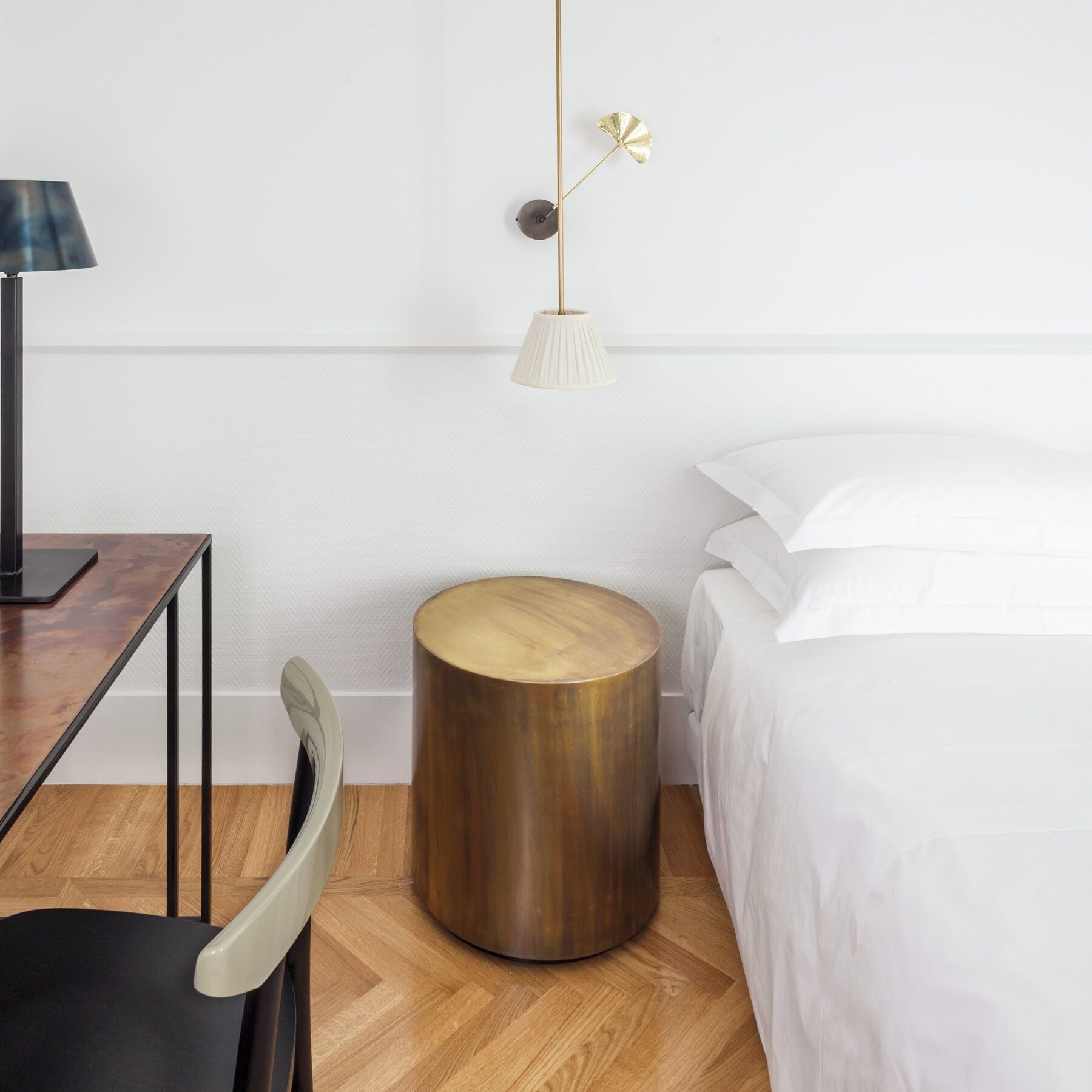 Une chambre à coucher au style minimaliste, avec une table d’appoint ronde en bois, un bureau avec une lampe et un lit recouvert de linge de lit blanc.