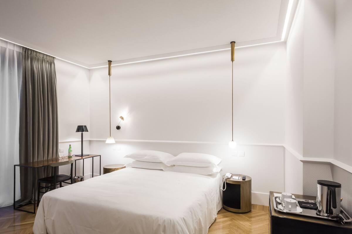 Una habitació d'hotel moderna amb un llit ben fet, una taula de nit amb una làmpada, una taula petita amb una cafetera i una cortina a la finestra.