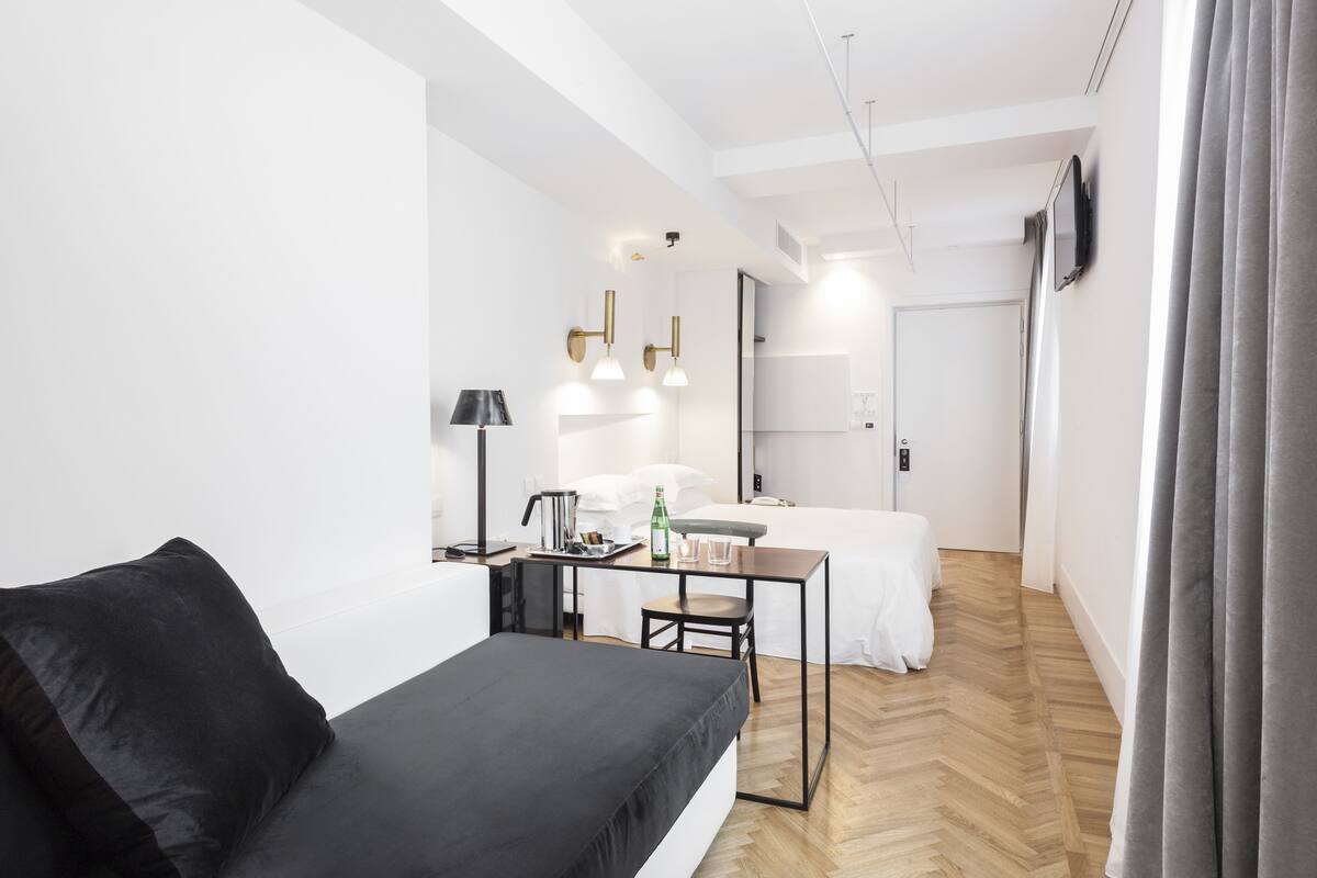 Habitació d'hotel moderna amb un sofà negre, una taula petita amb una làmpada, un llit i un mirall.