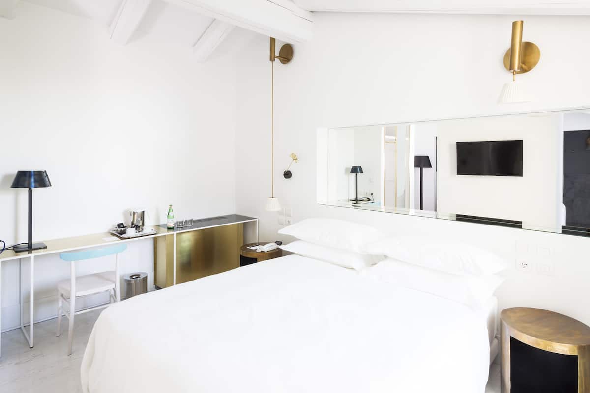 Habitació d'hotel amb un llit gran, una taula de treball amb una cadira, un tamboret petit, un mirall, una televisió i una làmpada fixa a la paret.