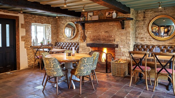 Un comedor rústico con pared de piedra, vigas de madera en el techo, chimenea y un espejo redondo.
