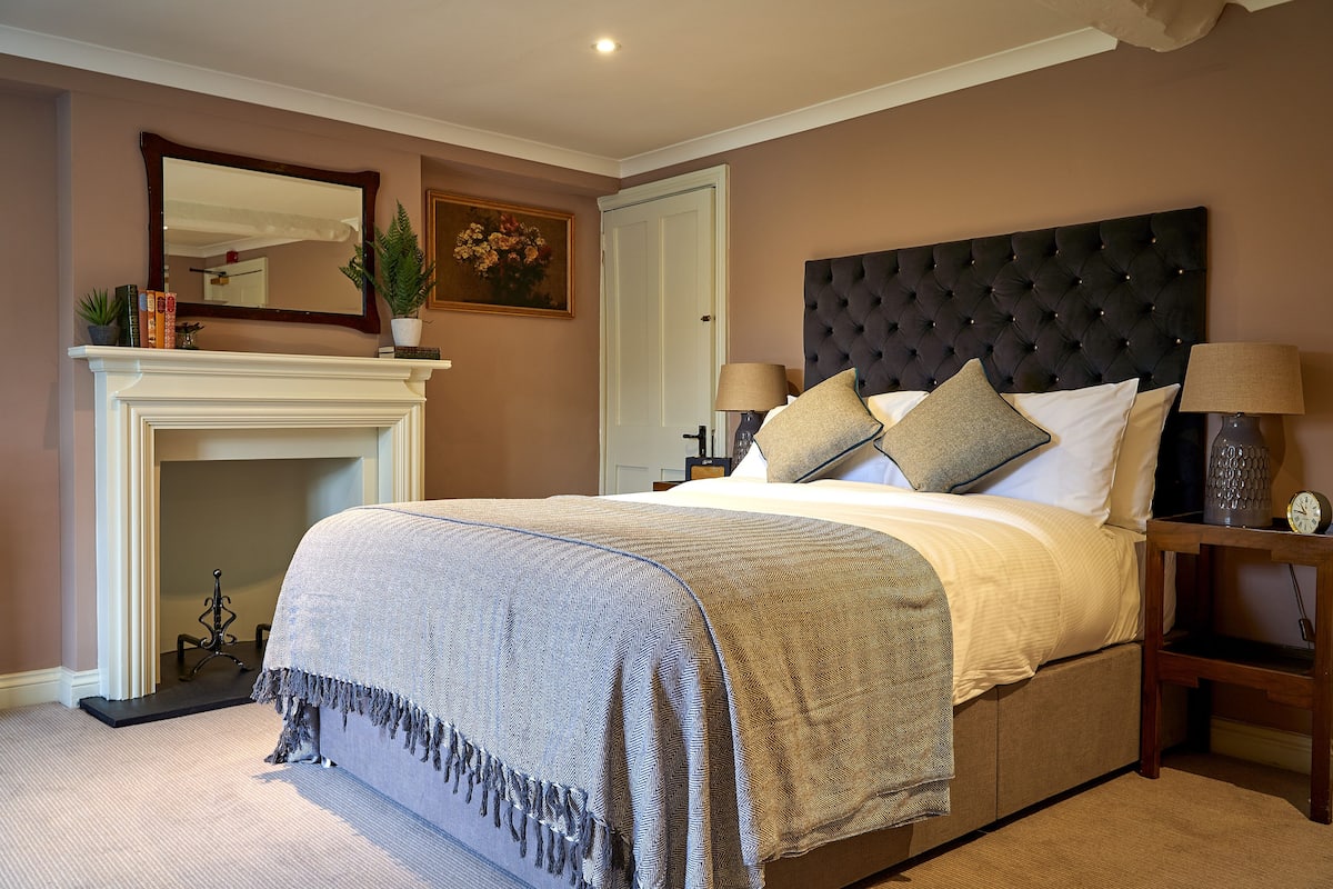 A bedroom with a large bed, a fireplace, a nightstand, and a mirror on the wall.